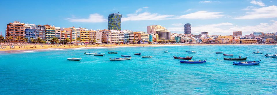 En solfylt strandpromenade med fargerike bygninger og små båter i det turkise havet.