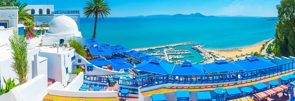 Panoramautsikt over en solfylt kystby med hvitkalkede bygninger, blå parasoller og asurblått hav.