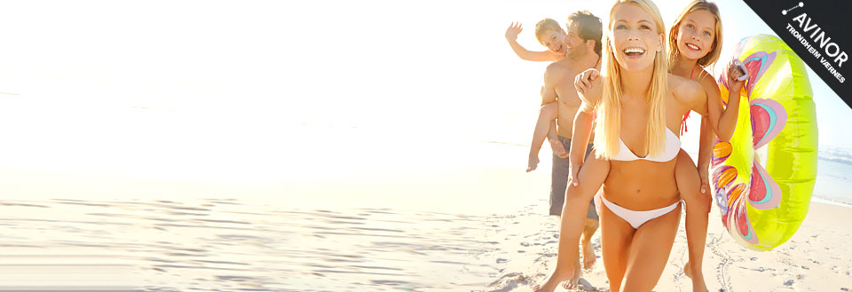 En familie koser seg i solen på stranda, barna leker, og en kvinne smiler med badetøy og badebøye.