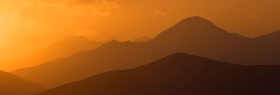 Bildet fremviser silhuetter av fjellkjeder mot en gyllen solnedgang fylt med varme farger.