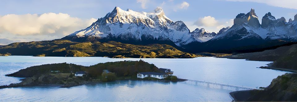 Bildet fremviser et fjellandskap med snødekte topper, en klar blå innsjø, og en liten øy med bygninger.