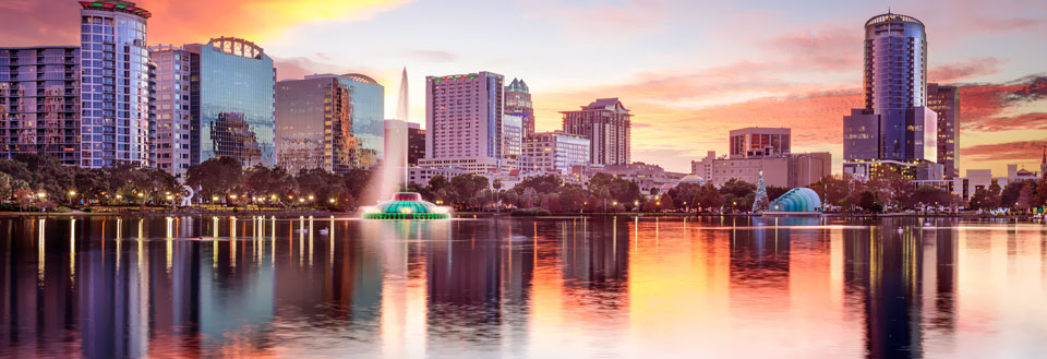 Orlandos skyline ved solnedgang med bygninger som speiler seg i et rolig vann.