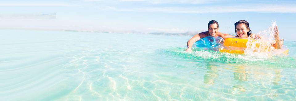 To personer ler og svømmer med oppblåsbare baderinge i krystallklart havvann.