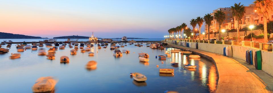Solnedgang over en havn på Malta med mange små båter. Vannet reflekterer den varme himmelen og kystlinjen er opplyst.