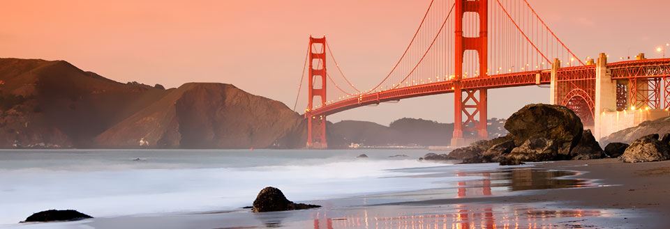 Solnedgangen bak Golden Gate Bridge med stille vann og strand forgrunnen.