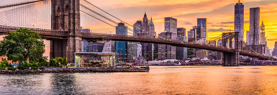 Bilde av en bro med  New Yorks skyline i bakgrunnen under solnedgang, refleksjon i vannet.