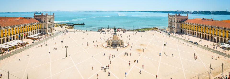 Panoramautsikt over et stort åpent torg i Lissabon med folk som går, omringet av klassiske bygninger, med havet i bakgrunnen.