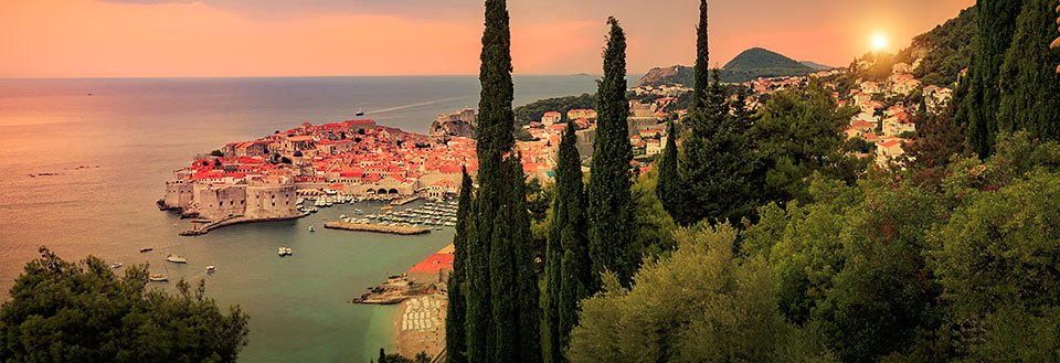 Bildet fremviser en sjarmerende solnedgang bak Dubrovnik omkranset av høye sypresser.