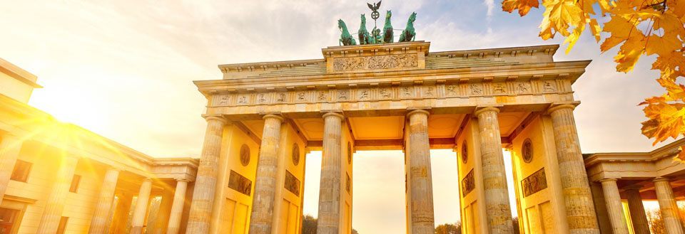 Bildet fremviser Brandenburger Tor i Berlin, opplyst av gyllen solnedgang, med høstløv foran.