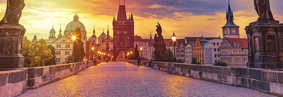 Solnedgangen kaster et gyldent lys over Karlsbroen i Prag med gamle tårn og statuer, bykonturene i det fjerne.