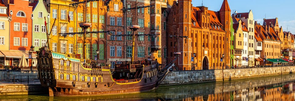 Fargerike bygninger speiler seg i vannet i Gdansk ved siden av en gammel seilskip i havnen.