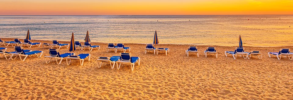 Bilde av tomme solsenger og parasoller på en sandstrand med stille hav i bakgrunnen under solnedgang.