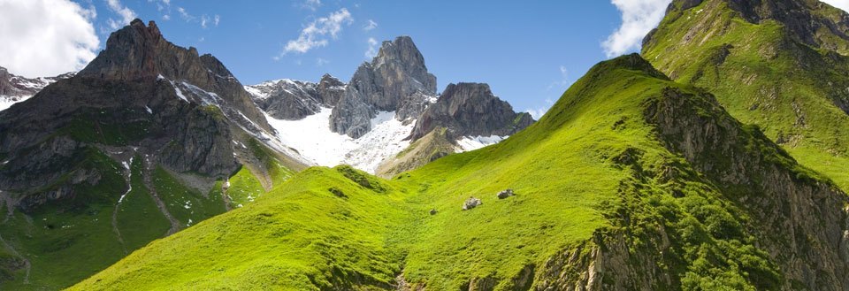 Frodig grønn dal med imponerende fjelltopper og snødekte topper i bakgrunnen under en klar blå himmel.