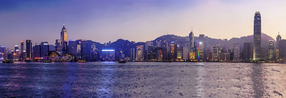 Skumring over Hong Kongs skyline med høye bygninger og belyste byggverk, speilet i vannet foran.