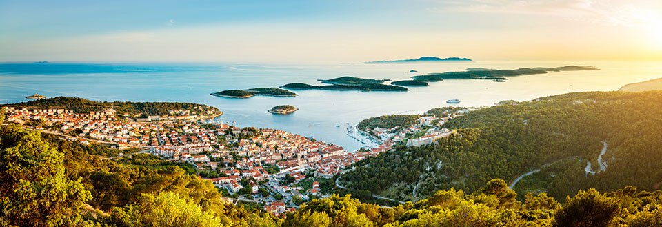 Panoramautsikt av en sjarmerende kystby i solnedgang, med grønne åser og øyer i det fjerne.