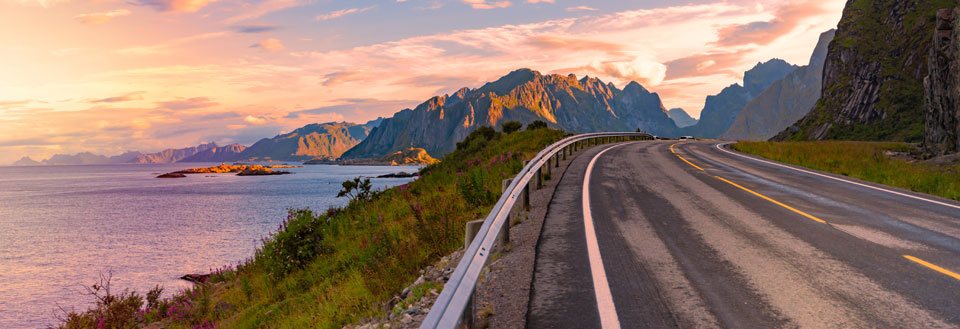 En tom vei snor seg gjennom et dramatisk landskap med fjell ved solnedgang.