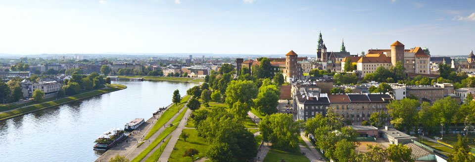 Panoramautsikt over en europeisk by med en elv, grøntarealer og historisk arkitektur under en klar himmel.