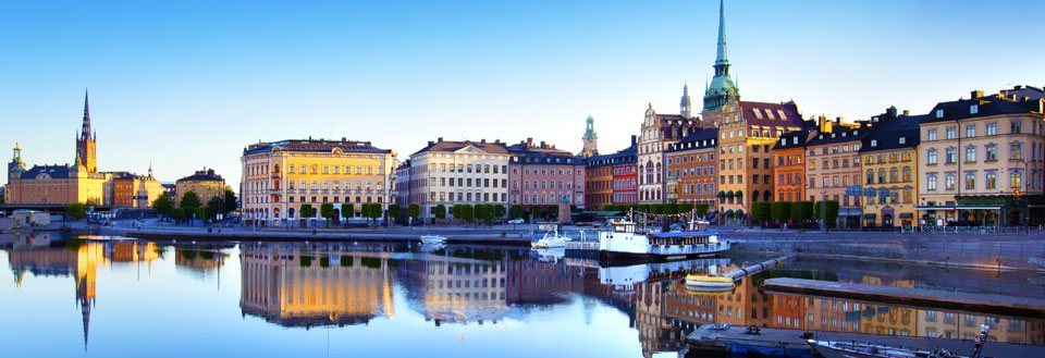 Panoramautsikt over en by ved vann i soloppgang med bygningers gjenspeiling i vannet.