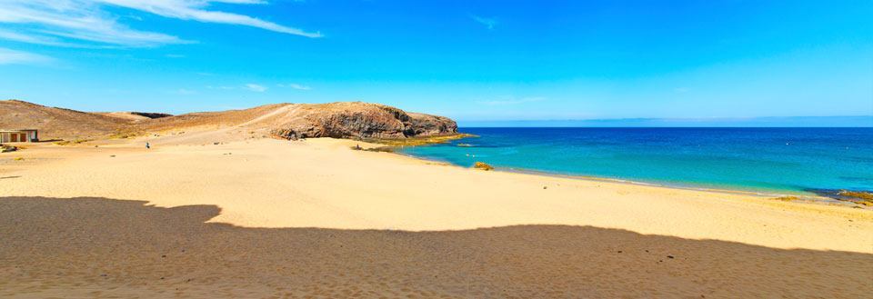 Bilde av en solfylt strand med gyllen sand, krystallklart blått hav og steinete klippe i bakgrunnen.