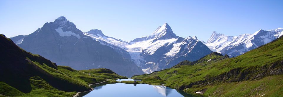 Bildet fremviser et pittoresk fjellterreng med en klar innsjø, flankert av grønne åser og snødekte topper.