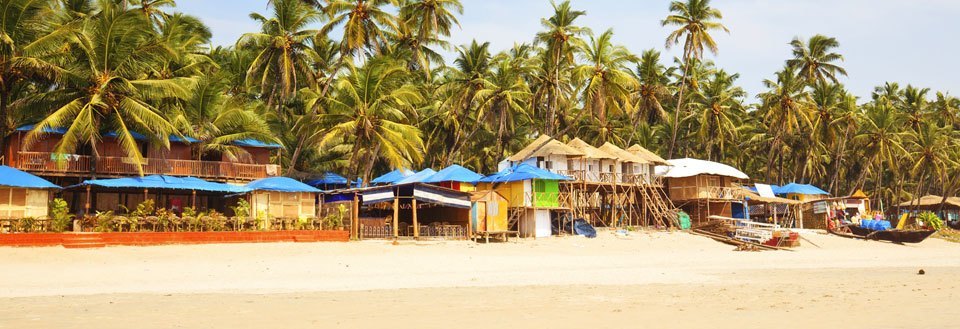 En tropisk strand i Goa med palmesus i bakgrunnen og små boder foran. Noen båter ligger på sanden.