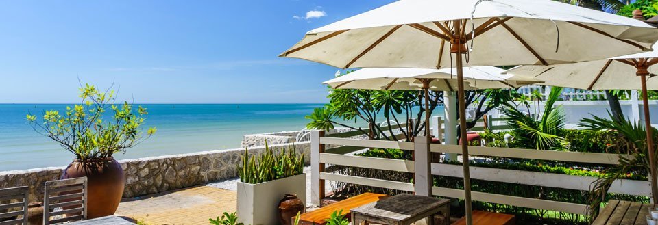 Terrasse med sjøutsikt, parasoller, stoler og frodige planter foran det klare blå havet.