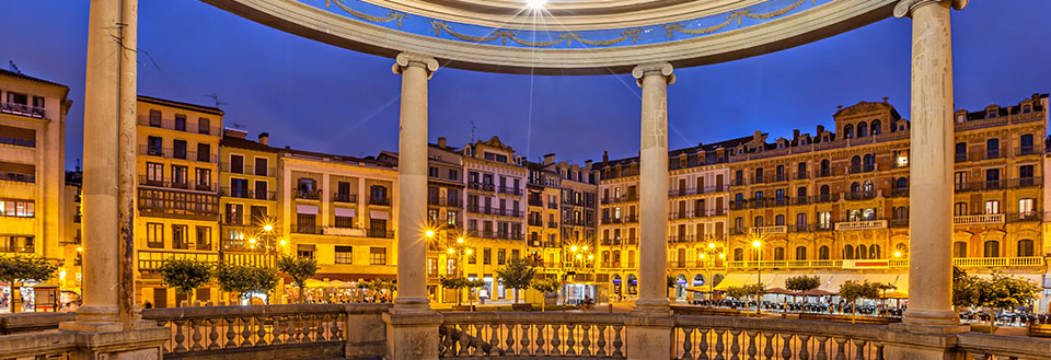 Et stemningsfullt kveldsbilde av et torg i Pamplona omgitt av klassiske bygninger opplyst av gatelys.