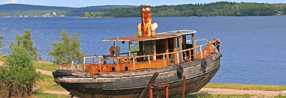 En rustet gammel båt på land med en skog og en innsjø i bakgrunnen.