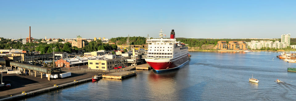 Havnefront med bygninger, et stort passasjerskip og små båter på en solrik dag.