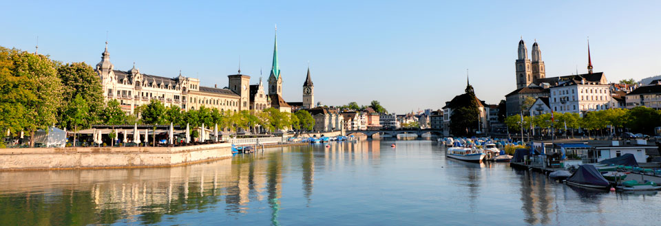 Et panoramabilde av en stille elv i en europeisk by med båter, bygninger og kirker.