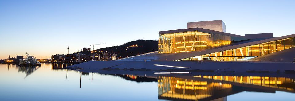 Moderne bygning reflekterer over stille vann ved skumringstid, med bylys i bakgrunnen.